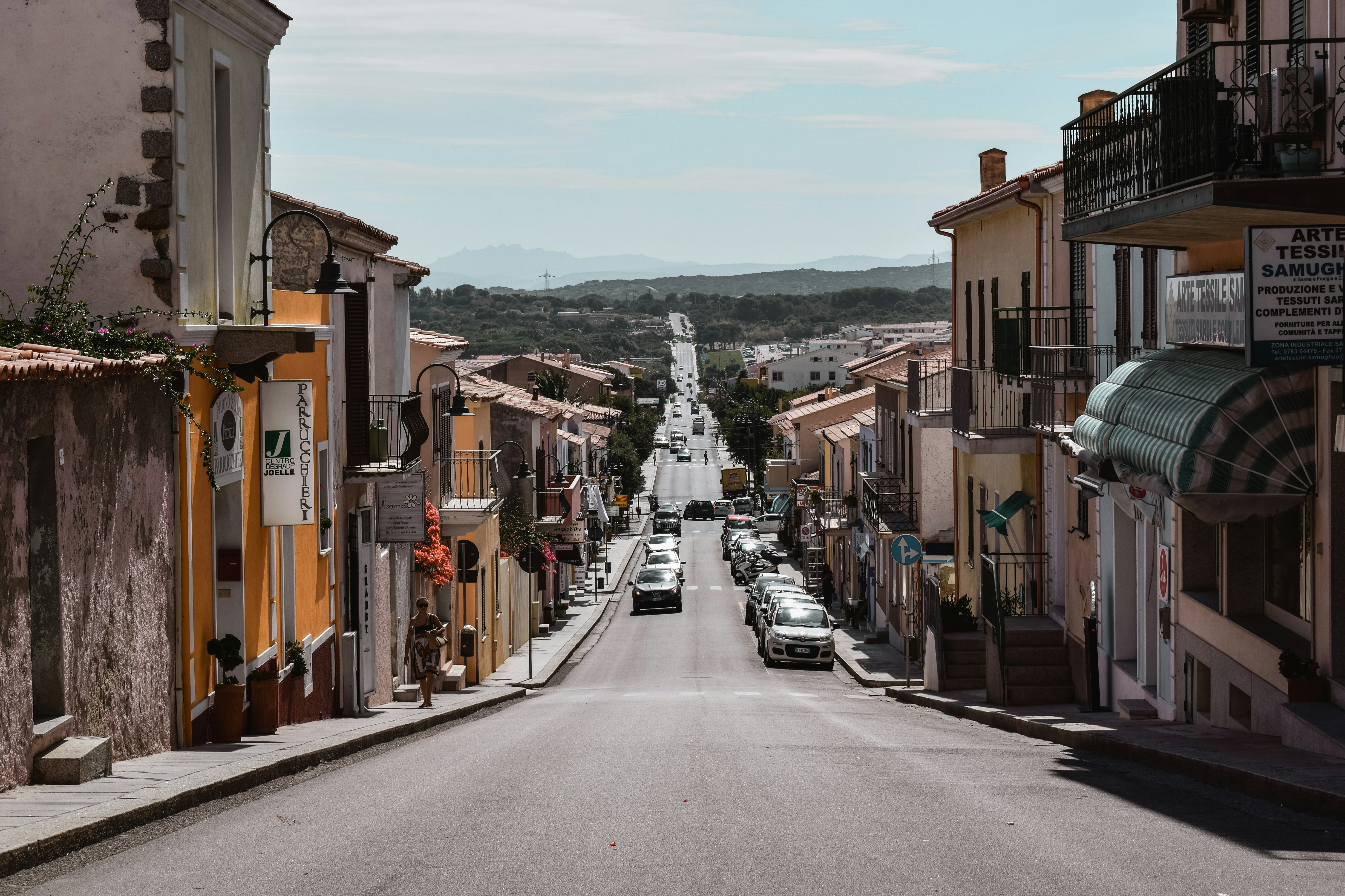 Cars parked along a quiet street in a Mediterranean town viewed downhill toward open countryside